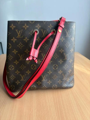 Louis Vuitton monogram axelväska - Snygg axelväska från Louis Vuitton i klassiskt brunt monogrammönster med detaljer och axelrem i rött läder. Väskan har guldfärgade metalldetaljer och dragsko upptill. Perfekt för dig som vill ha en ikonisk och trendig accessoar.