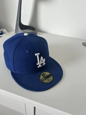 Blå LA New Era 59FIFTY keps - Snygg blå keps från New Era med LA-logga broderad i vitt framtill. Klassisk 59FIFTY-modell med platt skärm och klistermärke kvar på skärmen. Ventilationshål och vit knapp på toppen. Perfekt för dig som gillar streetwear och sportig stil.