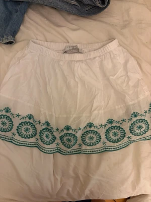Vit kortkjol med grön brodyr - Söt vit kortkjol från Primark med elastisk midja och detaljerad turkos brodyr längs nederkanten. Kjolen är gjord i ett tunt och luftigt bomullsmaterial, perfekt för varma dagar. Brodyren ger kjolen en unik och somrig känsla.