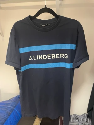 Svart t-shirt från J.Lindeberg - Snygg svart t-shirt från J.Lindeberg med två blå ränder och vit logga framtill. Klassisk rund halsringning och korta ärmar. Perfekt för en stilren och avslappnad look. Knappt använd 