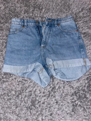 Blå denimshorts med uppvik - Klassiska blå jeansshorts i denim med hög midja och uppvikta benslut. Shortsen har fem fickor, bälteshällor och stängs med knapp och dragkedja. Perfekta för varma dagar och enkel att matcha med olika stilar.
