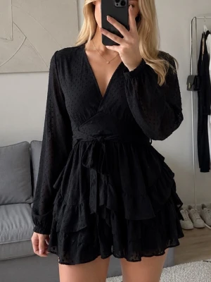 Svart omlott känning med volanger - Supersnygg svart jumpsuit med omlott-topp, långa ärmar och volangdetaljer på shortsdelen. Jumpsuiten har knyt i midjan och en lätt struktur i tyget som ger en extra touch. Perfekt för dig som vill sticka ut med stilren look.