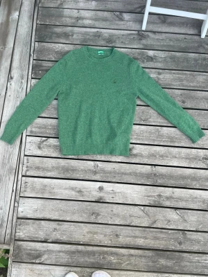 Grön stickad tröja i ull från united colors of benetton - En stilren grön stickad tröja med rund hals och långa ärmar. Tröjan har ribbstickade muddar vid ärmslut och nederkant. Perfekt för lager-på-lager och enkel att matcha med jeans eller kjol.