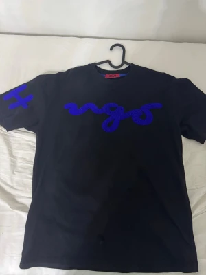 Tshirt - Svart t-shirt  med blå broderad logga framtill och blå detalj på ärmen. Klassisk passform och rund halsringning. Materialet är mjuk bomull som känns skönt mot huden. Perfekt för en stilren och modern look. L men passar mer som M