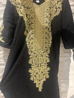 Svart klänning med guld broderi - Svart klänning med imponerande guldigt broderi längs halsringningen och framtill. Klänningen har lång ärm och ett vackert, detaljerat mönster som ger en lyxig känsla. Perfekt för dig som vill sticka ut med elegans.