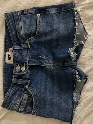 Low waist mini shorts från lager 157 - Säljer ett par blå jeansshorts från 157 med råa, fransiga kanter och dubbla knappar i midjan. Klassisk femficksmodell i denim med slitna detaljer för en avslappnad look. Perfekta till sommaren!