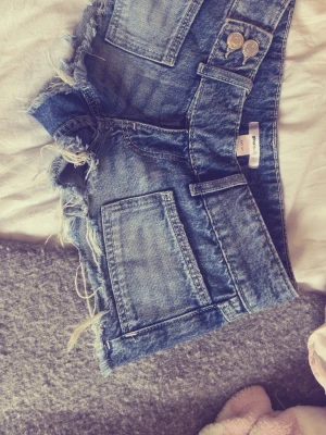Blå low waisted jeansshorts från Gina Tricot - Säljer ett par blå low waist jeansshorts med fransig kant och stora fickor både fram och bak. Shortsen har två silvriga knappar i midjan och en klassisk denimlook. Perfekta för varma dagar och en avslappnad stil.