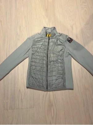Ljusgrå jacka från parajumper  - Ljusgrå vindjacka från parajumper med quiltade partier fram och bak samt släta ärmar. Jackan har hög krage, hel dragkedja och en bröstficka med dragkedja. Patch med logga på vänster ärm och diskret ficka bak. Materialet ser ut att vara syntet.