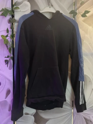 Svart hoodie med blå detaljer Adidas - Svart hoodie från Adidas med blå paneler längs axlar och ärmar. Klassisk huva och stor magficka framtill. Adidas-logga på bröstet och vita ränder på ena ärmen. Perfekt för en sportig och avslappnad stil.