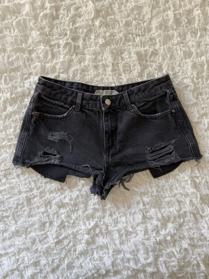 Svarta lågmidjade jeansshorts  - Snygga mörkgrå/svarta jeansshorts med rå fåll och slitna detaljer framtill och baktill. Perfekta till sommaren och ger en cool, edgy look. I fint skick utan anmärkningar utöver den avsiktligt slitna designen. 💕 Midjemåttet är ungefär 36cm (alla mått finns i bilderna).