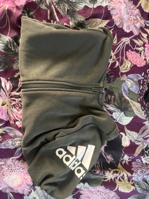 Mörkgrön hoodie från Adidas - Snygg mörkgrön hoodie från Adidas med dragkedja framtill och stor vit Adidas-logga på ena ärmen. Hoodien har en klassisk huva och är perfekt för en avslappnad streetwear-look. Tillverkad i mjukt material för extra komfort.