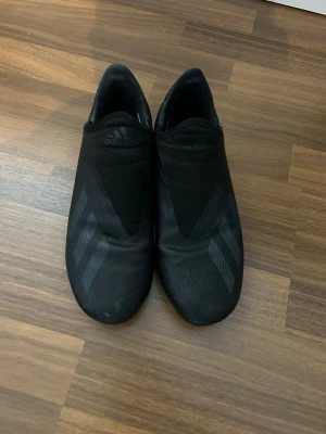 Svarta Adidas fotbollsskor utan snörning - Säljer ett par helsvarta fotbollsskor från Adidas med stilrena, diskreta ränder på sidorna. 