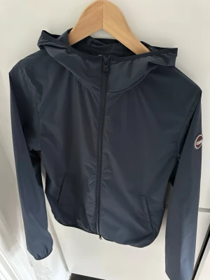 Mörkblå vindjacka från Colmar - Snygg mörkblå vindjacka från Colmar Originals med huva och dragkedja framtill. Jackan har två fickor och en diskret logga på ärmen. Perfekt för blåsiga dagar och enkel att matcha med olika outfits.