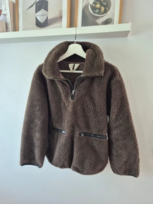 Brun teddyjacka från ARKET  - Mysig brun teddyjacka från Arket med halv dragkedja framtill och två svarta dragkedjefickor. Jackan har hög krage och är tillverkad i fluffigt teddy-material som håller dig varm under kyliga dagar. Perfekt för en trendig och avslappnad look.