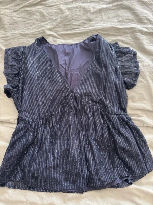 Glittrig svart blus med volangärm - Snygg svart blus med glittriga ränder och v-ringning. Toppen har korta volangärmar och en lätt utsvängd passform nedtill. Perfekt för dig som vill sticka ut lite extra med en glittrig look.