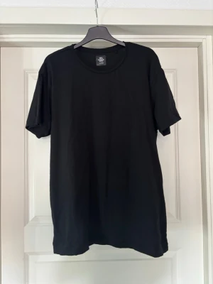 Svart basic t-shirt i bomull - Enkel svart t-shirt med rund hals och korta ärmar. Tillverkad i mjuk bomull och har en normal passform. Perfekt att matcha med jeans eller shorts för en clean och stilren look.
