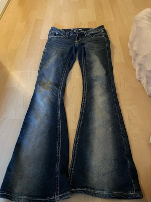 Mörkblå utsvängda jeans med broderi - Säljer ett par mörkblå jeans med kraftigt utsvängda ben och slitningar. Jeansen har snygga broderade detaljer och dekorationer på bakfickorna. Tydliga kontrastsömmar och klassisk femficksmodell. Perfekta för dig som gillar retrostil!