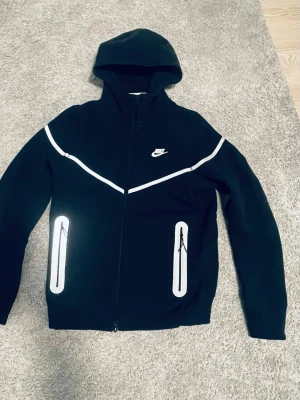 Svart Nike tech hoodie med dragkedja - Använd en gång! Svart hoodie från Nike med hel dragkedja, reflex/silver logga på bröstet och silver/reflex detaljer längs bröstet och fickorna. Utrustad med huva och två fickor framtill. Perfekt för en sportig och avslappnad stil. Passar typ för 175.