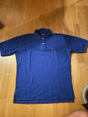 Blå pikétröja från Yves Saint Laurent - Snygg blå pikétröja från Yves Saint Laurent i 100% bomull. Klassisk krage med två knappar och diskret logga broderad på bröstet. Perfekt för en stilren och avslappnad look.