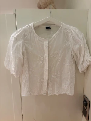 Vit blus med broderi från Gina Tricot - Söt vit blus från Gina Tricot med puffiga trekvartsärmar och fina blommiga broderier. Blusen har rund halsringning och knäppning framtill. Tillverkad i ett lätt och luftigt tyg som ger en romantisk känsla.