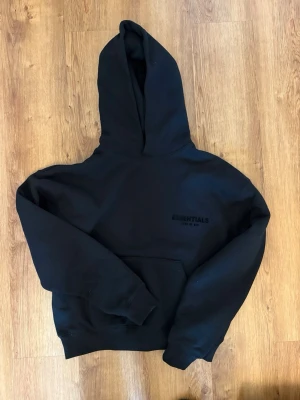 Svart Essentials Fear of God hoodie - Säljer en svart hoodie från Essentials Fear of God med broderad logga på bröstet. Hoodien har en stor huva och magficka framtill. Tillverkad i mjukt bomullsmaterial och har en avslappnad passform som är perfekt för en chill look. Storlek XXS men passar mer som xs-s. Knappt använd och finns även byxor 