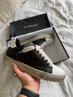 Numeris Atelier Waxed - Storlek 45 - Snygga sneakers från Numeris i svart och vitt läder med breda beige skosnören och grov sula. Skon har en låg profil och kontrasterande vit häl och tåparti. Perfekt för dig som vill ha en stilren men ändå unik sneaker till vardagslooken.