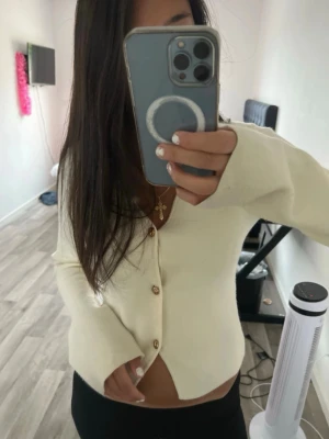 Krämvit kofta med träknappar - Snygg krämvit kofta med v-ringning och träfärgade knappar framtill. Modellen är figurnära med långa ärmar och mjukt material som känns skönt mot huden. Perfekt att styla till både jeans och kjol för en clean look.