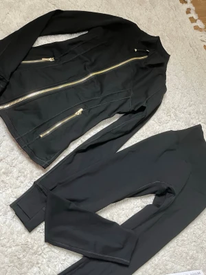 Svart träningsset Lululemon  - Snyggt svart träningsset med jacka och tights. Jackan har figurnära passform, ståkrage och guldiga dragkedjor framtill och på fickorna. Tightsen är höga i midjan och har en slimmad look. Perfekt för gymmet eller löprundan.