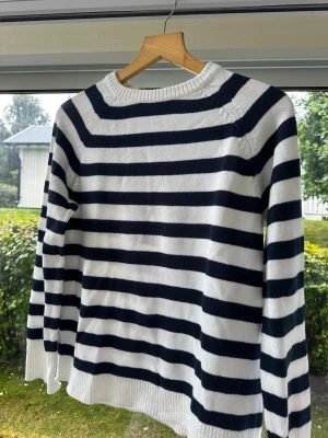 Randig stickad tröja i marinblått och vitt - Snygg stickad tröja med klassiska marinblå och vita horisontella ränder. Tröjan har rund halsringning, långa ärmar och ribbstickade muddar vid ärmslut och nederkant. Perfekt för sommarkvällar, våren eller hösten. Storlek S men oversized.