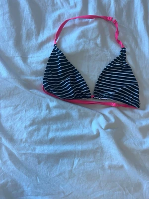 Randig bikiniöverdel med rosa band - Snygg bikiniöverdel med svart och vitt randigt tyg och starkt rosa band för knyt i nacken och ryggen. Klassisk trekantsmodell! Aldrig använd!