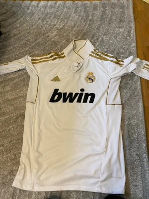 Real Madrid långärmad matchtröja - Vit långärmad Real Madrid matchtröja från Adidas med guldfärgade detaljer och klubbmärke på bröstet. Tröjan har svart Bwin-tryck på framsidan och klassiska Adidas-ränder på axlarna. Tillverkad i ett lätt och funktionellt material.