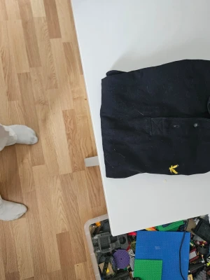 Svart skjorta med gul broderad logga - Snygg svart pike med en gul broderad logga på bröstet. Skjortan har klassisk krage och knäppning framtill. Perfekt för dig som gillar stilrena plagg med en liten detalj. Materialet känns mjukt och bekvämt.