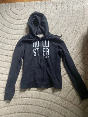 Mörkblå Hollister hoodie med dragkedja - Snygg mörkblå hoodie från Hollister med vit broderad logga på bröstet och texten 'SURF'. Hoodien har dragkedja framtill, två fickor och justerbar huva med snören. Perfekt för en avslappnad och cool stil. Står strl L men liten i strl så passar xs/s❤️