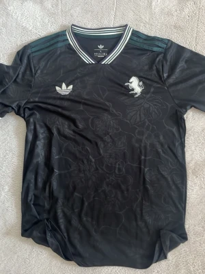 Svart Adidas sporttröja med mönster - Snygg svart sporttröja från Adidas med diskret blommönster i tyget. Tröjan har vita Adidas-loggan och ett vitt lejon på bröstet samt randiga detaljer i grönt och vitt på kragen och axlarna. Perfekt för träning eller streetwear.