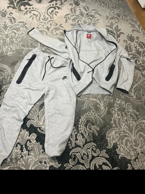 Nike Tech Tracksuit - Snyggt grått mjukisset från Nike med svarta dragkedjor och detaljer. Setet består av en ziptröja och matchande byxor med dragsko i midjan. Perfekt för chill eller träning. Materialet är mjukt och stretchigt för maximal komfort. Nike Tech Tracksuit, använd 1-2 gånger annars helt ny i skick utan prislapp! Pris kan diskuteras via dm hör av er! Storlek L men passar M!