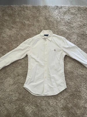  polo Ralph Lauren Oxford - Klassisk vit skjorta med lång ärm och knappar framtill. Skjortan har en broderad logga på bröstet och är tillverkad i bomull. Perfekt till både jeans och chinos för en clean look.