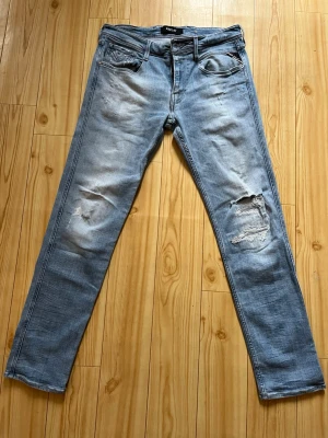 Replay jeans - Säljer ett par ljusblå jeans med raka ben och slitningar på både lår och knä. Jeansen har klassisk femficksmodell och tydliga hål på höger knä för en trendig, avslappnad look. Perfekta för dig som gillar streetstyle och en casual vibe.