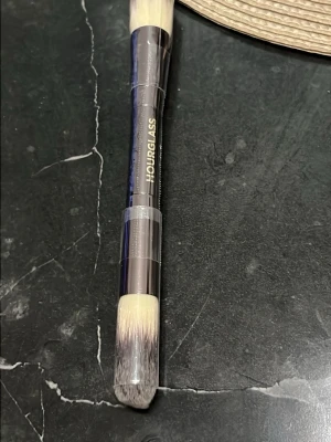Hourglass dubbelsidig sminkborste - Snygg dubbelsidig sminkborste från Hourglass med metalliskt handtag och täta, mjuka syntetiska borst i beige och grå toner. Perfekt för applicering av foundation, concealer eller contour. Smidig att ha i necessären och enkel att använda för olika makeupsteg.