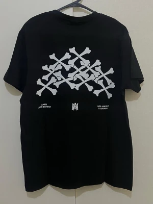 Svart AMIRI t-shirt med benkors - Säljer en svart t-shirt från AMIRI med tryck av vita benkors på ryggen och AMIRI-logga framtill. T-shirten har rund hals och korta ärmar. Materialet är mjuk bomull och passformen är normal. Perfekt för dig som gillar streetwear och vill sticka ut.