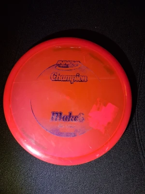 Röd discgolf-disc Innova Champion mako 3 - En röd discgolf-disc från Innova, modell Champion Mako3. Tillverkad i plast med tryckt text i blått. Perfekt för dig som gillar discgolf och vill ha en stabil midrange-disc som går spik rakt!