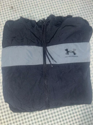Svart vindjacka från Under Armour - Snygg svart vindjacka från Under Armour med grått bröstparti och logga på vänster sida. Jackan har huva, dragkedja framtill och är perfekt för blåsiga dagar. Tillverkad i lätt polyester som andas och ger en sportig look.
