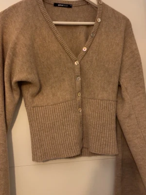 Beige kofta från Gina Tricot - Mysig beige kofta från Gina Tricot med v-ringning och knappar framtill. Koftan har ribbstickad nederdel och långa ärmar. Perfekt att slänga över en topp för en avslappnad look.