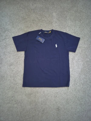 Polo Ralph Lauren - Snygg mörkblå t-shirt från Polo Ralph Lauren med klassisk vit logga broderad på bröstet. T-shirten har rund hals och korta ärmar, perfekt för en clean och stilren look. Tillverkad i mjuk bomull för skön komfort.