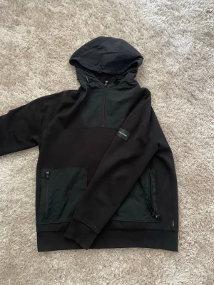 Calvin Klein Hoodie - Snygg svart hoodie med huva och halv dragkedja framtill. Har två stora fickor med dragkedja och en patch på ärmen. Materialet är en mix av bomull och nylonpaneler som ger en cool kontrast. Perfekt för en avslappnad streetwear-look.