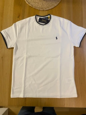 Vit t-shirt från Polo Ralph Lauren - Klassisk vit t-shirt från Polo Ralph Lauren med svart logga broderad på bröstet. T-shirten har svarta detaljer vid krage och ärmslut samt en normal passform. Perfekt till jeans eller shorts för en clean och stilren look.