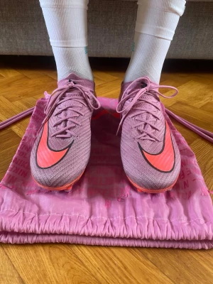 Rosa Nike fotbollsskor med dobbar - Säljer ett par snygga fotbollsskor från Nike i rosa med svarta och neonrosa detaljer. Skorna har snörning, låg profil och mönstrad ovandel i syntetmaterial. Dobbarna är utformade för bra grepp på gräsplan. Perfekta för dig som vill sticka ut på planen!