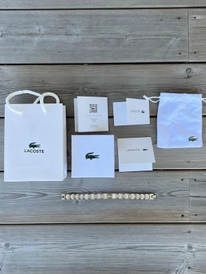 Silverfärgat armband från Lacoste - Snyggt armband i silverfärgad metall från Lacoste med den klassiska krokodilloggan ingraverad på mitten. Länkarna har en stilren och modern design med diskreta detaljer. Perfekt accessoar för dig som gillar en clean och sportig look. Längden går att anpassa, man kan ta bort länkar. Aldrig använt.