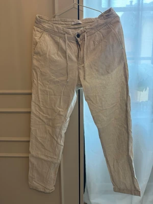 Beige linnebyxor från John Henric - Snygga beige linnebyxor från John Henric med avslappnad passform och raka ben. Byxorna har snedfickor fram, knappgylf och dragsko i midjan för extra komfort. Perfekta för en chill och stilren look under varma dagar.