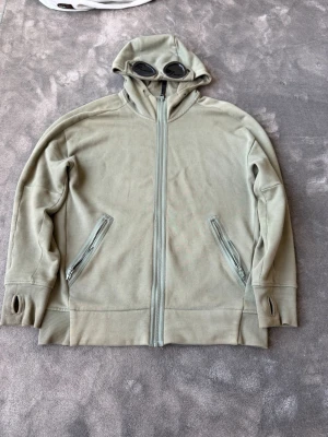 Beige hoodie med glasögon från C.P. Company - Säljer en beige hoodie från C.P. Company med hel dragkedja, två fickor med dragkedja och ikoniska glasögon i huvan. Tröjan har tumhål i ärmsluten och är gjord i mjukt bomullsmaterial. Perfekt för dig som vill sticka ut med en unik detalj!