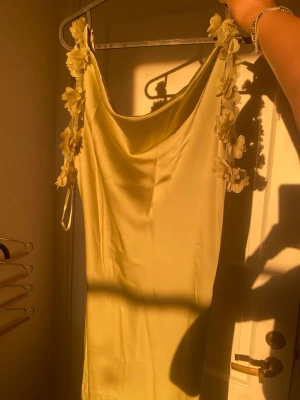 Ljusgrön klänning med blommiga axelband - Supersöt ljusgul klänning från Vero Moda med tunna axelband dekorerade med 3D-blommor. Klänningen har en mjukt draperad urringning och är i ett slätt, glansigt material som ger en elegant look. Perfekt för sommarens alla tillfällen! (Den är helt ny med prislapp, aldrig använd)